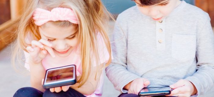 Zwei Kinder, ein Junge und ein Mädchen, sitzen nebeneinander und haben jeweils ein Handy in der Hand. Ihre Blicke liegen auf den Mobilgeräten vor ihnen.